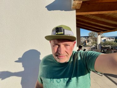 Mann in türkisem Shirt hält Insta360 Go Ultra in die Kamera, Selfie-Aufnahme im Freien mit Vegetation und Himmel