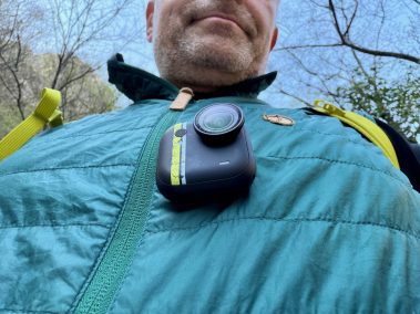 Mann in türkiser Jacke mit Insta360 Go Ultra am Bruststrapez, Selfie-Perspektive von unten, Himmel im Hintergrund