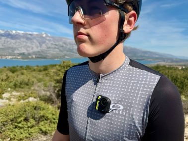 Rennradfahrer in grauem Trikot trägt Insta360 Go Ultra am Bruststrapez, Küstenlandschaft und Meer im Hintergrund