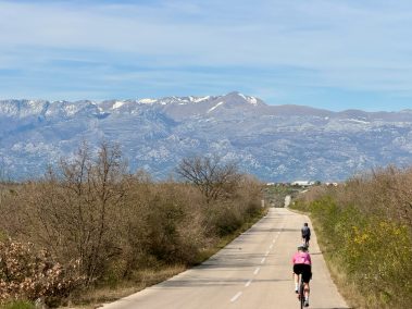 Langer gerader Radweg durch mediterrane Landschaft mit Olivenbäumen, Rennradfahrerin in der Ferne – Insta360 Go Ultra