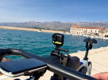Insta360 Go Ultra Kamera am Fahrradlenker montiert, Hafenort mit Booten und Bergen im Hintergrund
