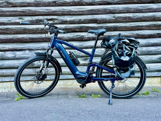 MapFour Roam blau mit Seitentasche vor rustikaler Holzwand – Touring Setup